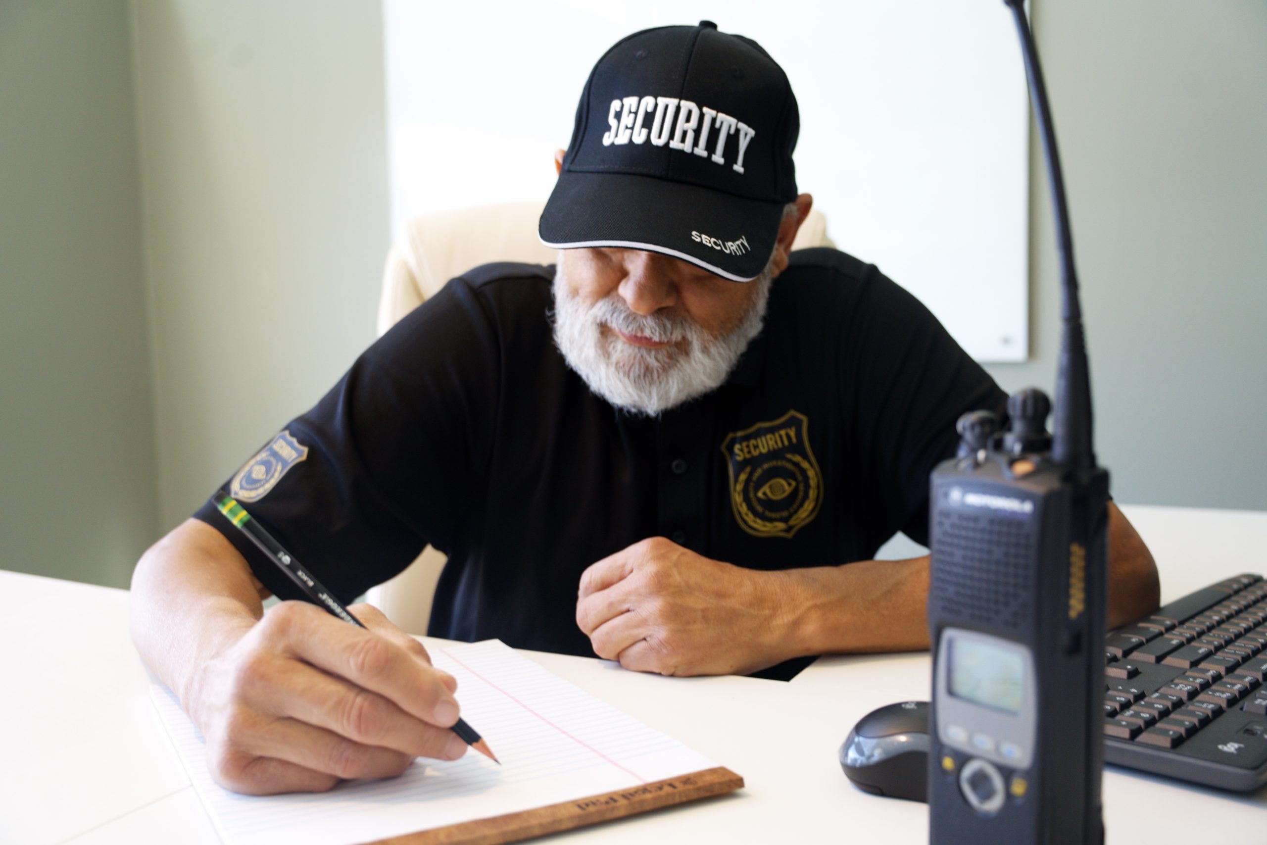 Security guard writing notes at a desk, wearing a "SECURITY" cap, with a two-way radio and computer mouse nearby, reflecting corporate security operations in Odessa.