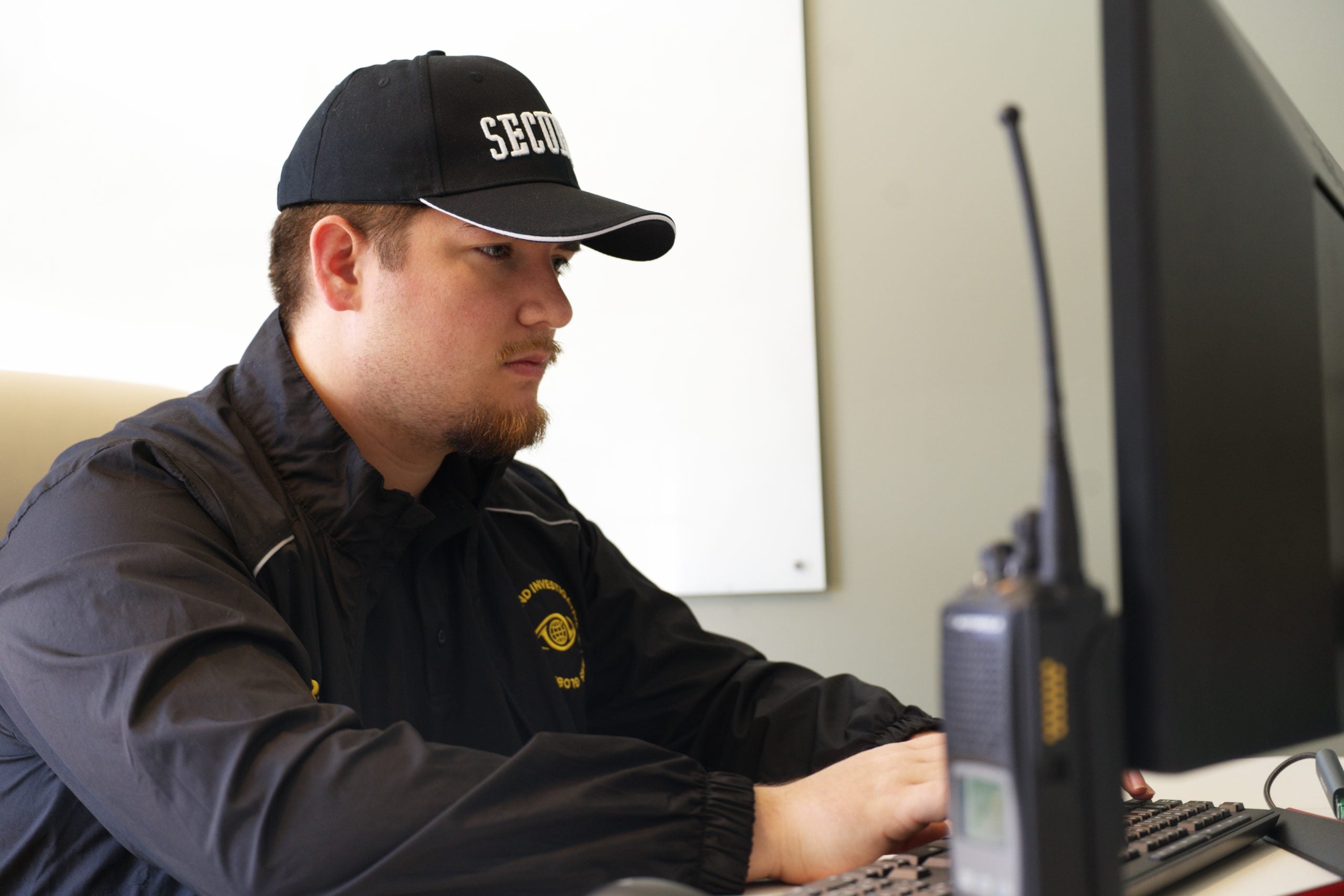 Security professional in a cap and jacket, focused on computer work, with communication equipment nearby, emphasizing reliability in security services.