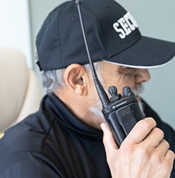 Security personnel using a radio communication device, wearing a "SECURITY" cap, emphasizing professional security services and monitoring.