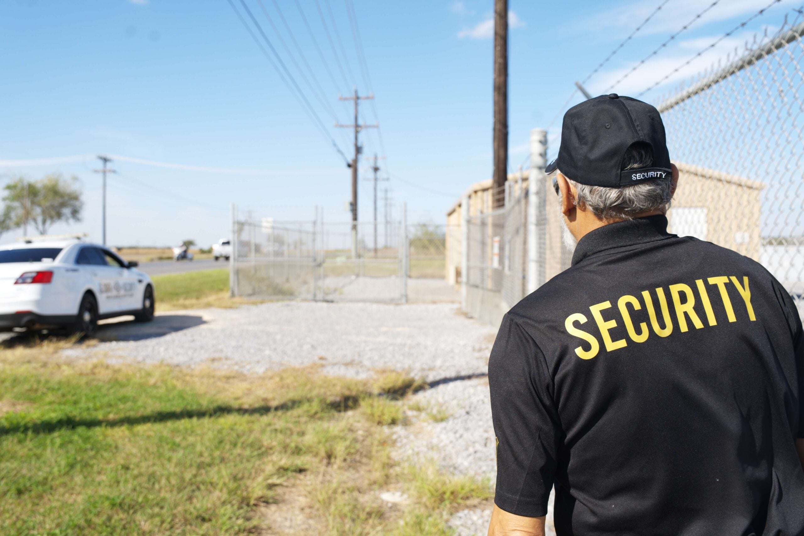 Security guard in uniform monitoring a secured area near a fence, with a patrol car visible, emphasizing professional security services in Houma, Louisiana.