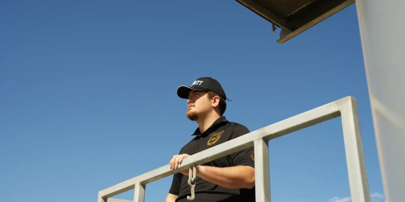 Security guard observing surroundings, wearing a branded cap and uniform, against a clear blue sky, reflecting executive security services.