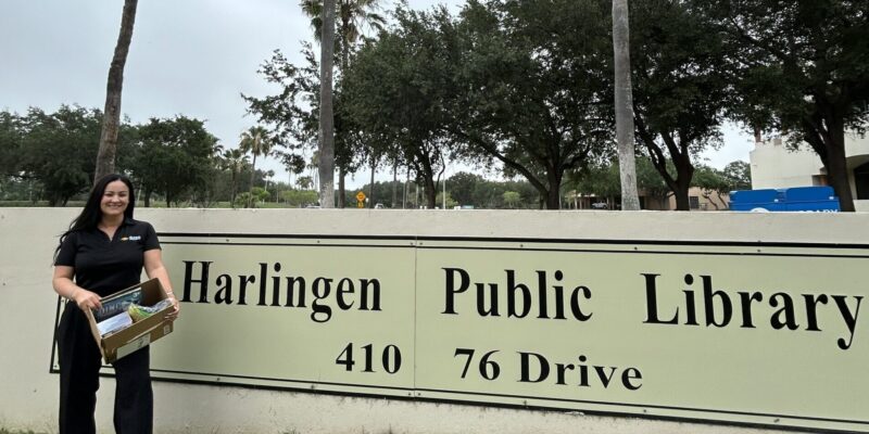 GSIS CFO Alva Salinas stands outside the Harlingen Public Library holding donated items from the library’s community wishlist near the library entrance sign.