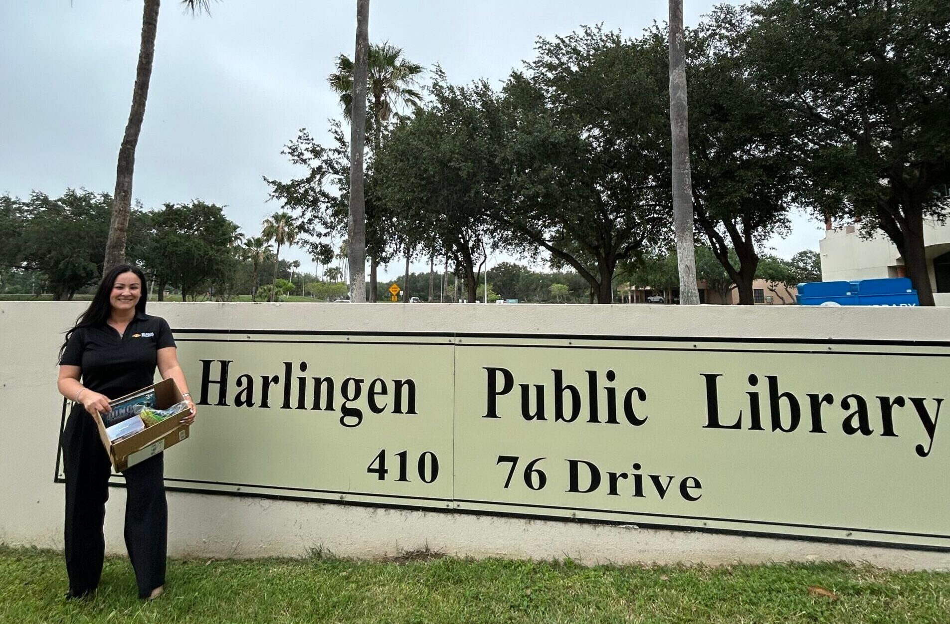 GSIS CFO Alva Salinas stands outside the Harlingen Public Library holding donated items from the library’s community wishlist near the library entrance sign.