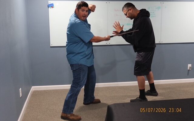 Security training instructor demonstrating baton handling and defensive techniques during a Level III security guard training class while a student practices a blocking stance indoors.