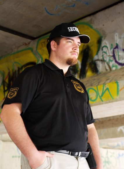 Armed security guard in black uniform with "SECURITY" cap, standing confidently under an overpass with graffiti, representing professional security services in Louisiana.