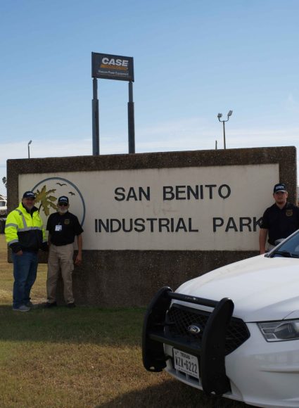 Security personnel standing beside a sign for San Benito Industrial Park, with a patrol car in the foreground, illustrating the importance of trained security in industrial settings.